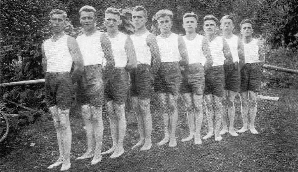 Schwarz-Weiß-Foto von 1919: Neun junge Turner in weißen Trikots und dunklen Hosen stehen barfuß nebeneinander auf einer Wiese.