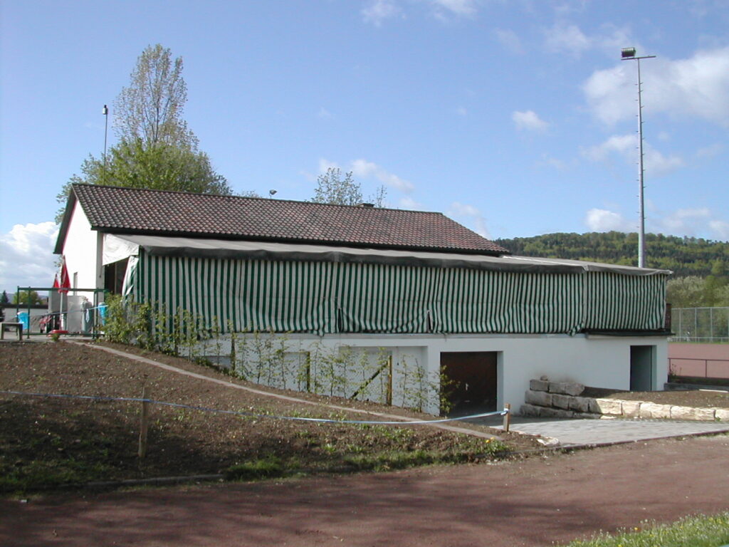 Das Vereinsheim nach der Erweiterung: weißes Gebäude mit grün-weiß gestreifter Markise am Sportplatz, daneben ein Flutlichtmast.