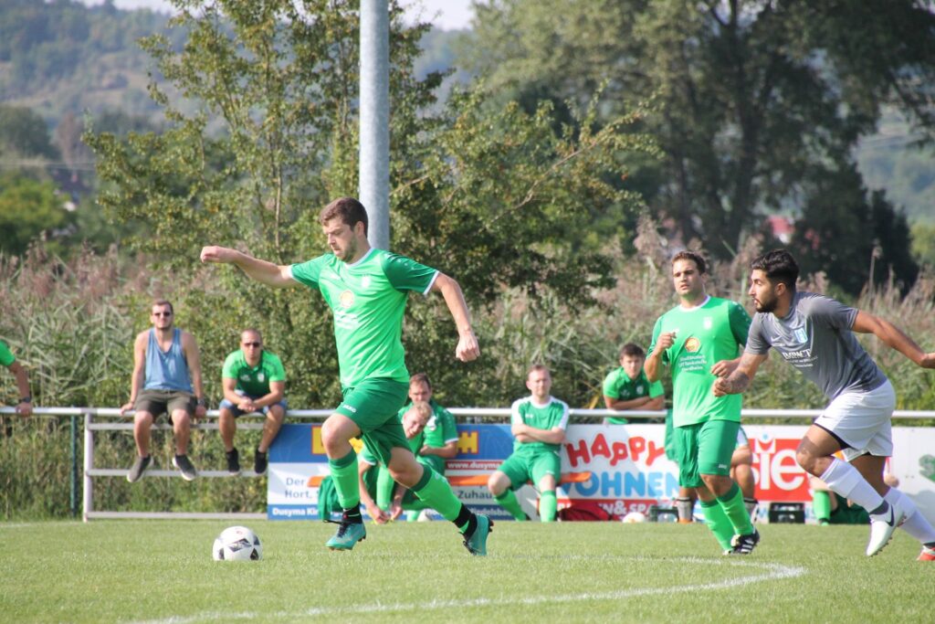 Mehrere Fußballspieler sind während eines Spiels auf dem Platz zu sehen; ein Spieler in grünem Trikot führt den Ball, während ein Gegenspieler in Grau auf ihn zuläuft und weitere Spieler sowie Zuschauer im Hintergrund stehen.