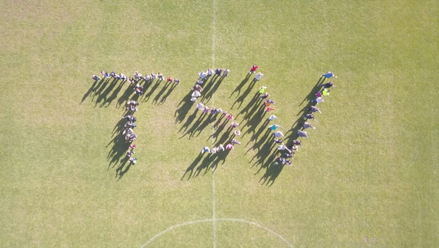 Luftaufnahme vom Sportplatz: Rund hundert Mitglieder stehen auf dem Rasen und bilden gemeinsam den Schriftzug TSV.