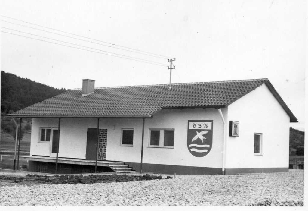 Das fertige Vereinsheim 1969: weißes Gebäude mit Ziegeldach, überdachter Terrasse und großem TSV-Wappen an der Wand.