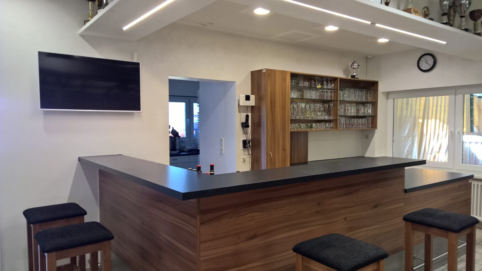 Theke mit dunkler Arbeitsplatte und Barhockern, dahinter Gläserregal, Durchgang zur Küche und ein Fernseher an der Wand