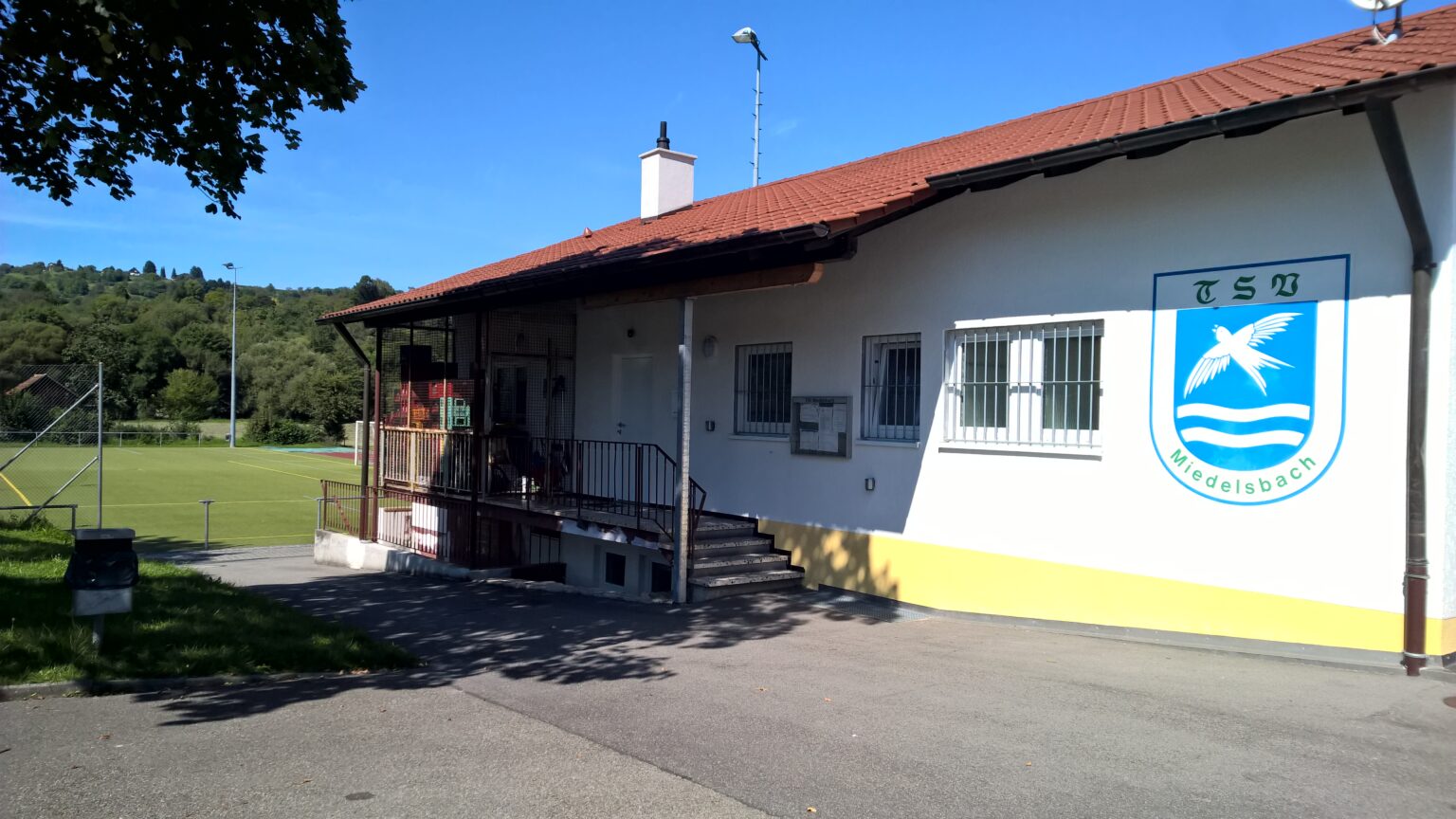 Vereinsheim des TSV Miedelsbach von außen: weißes Gebäude mit rotem Ziegeldach und großem TSV-Wappen an der Wand, daneben der Kunstrasenplatz